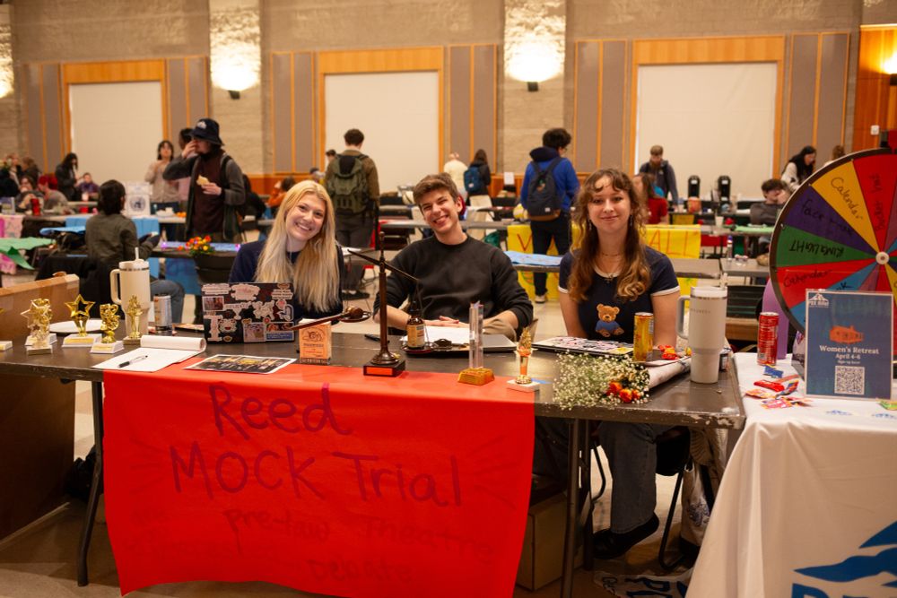 Three Reed students sitting at a table. There is a red piece of paper in front of the table which reads, "Reed Mock Trial."