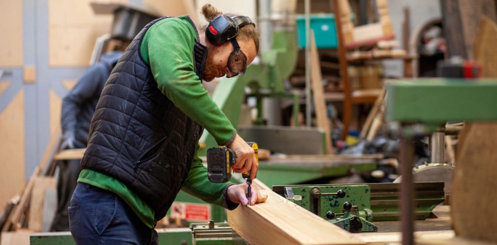A contractor, Boris Afinogenov, works on a piece of timber, drilling into it. He is wearing safety goggles! 