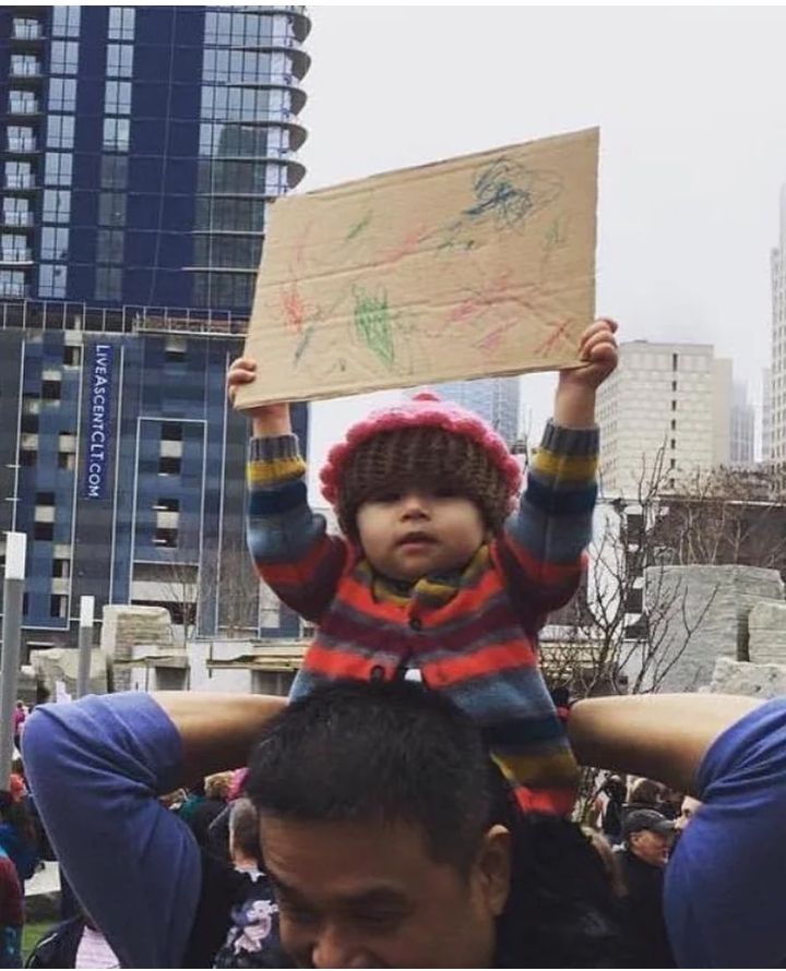 Photo of baby at a protest holding up a cardboard sign with crayon scribbles