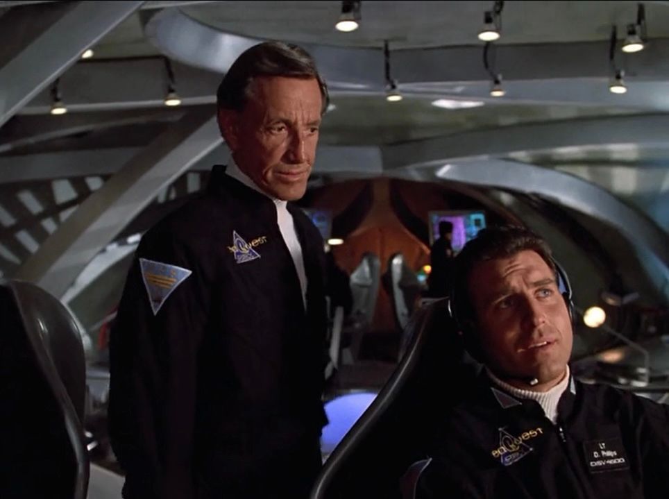 Two men wearing black jumpsuits. One is seated and wearing a headset, the other stands over the seated man’s right shoulder. They are on the bridge of an advanced submarine.