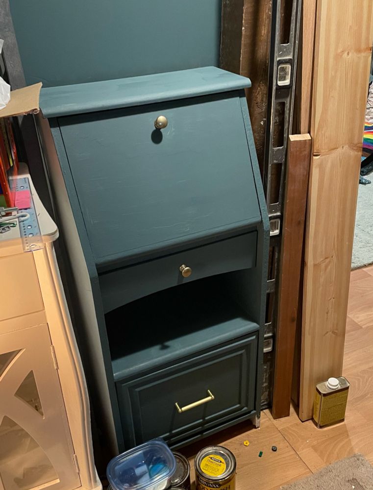 A small secretary desk is tucked into a wall next to a doorway. There are construction materials to its right and paint and stain cans on the floor. 
The desk and the walls are painted the same color: Sherwin Williams Riverway, and the desk has two brass knobs and a brass handle.