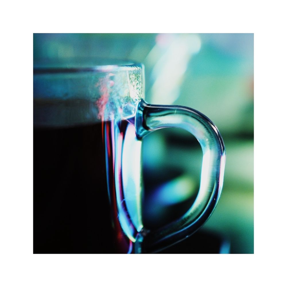 A portrait of a clear glass coffee mug reflecting greens, blues, purples, and highlights with a bokeh background.