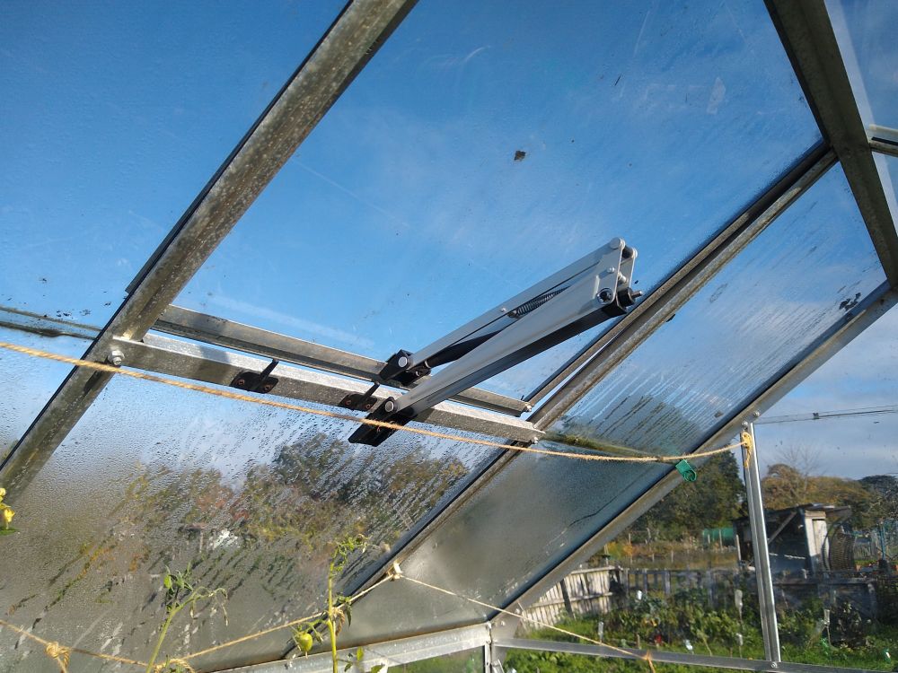 Greenhouse automatic roof vent just cracking open, with a beautiful blue sky visible behind it.
