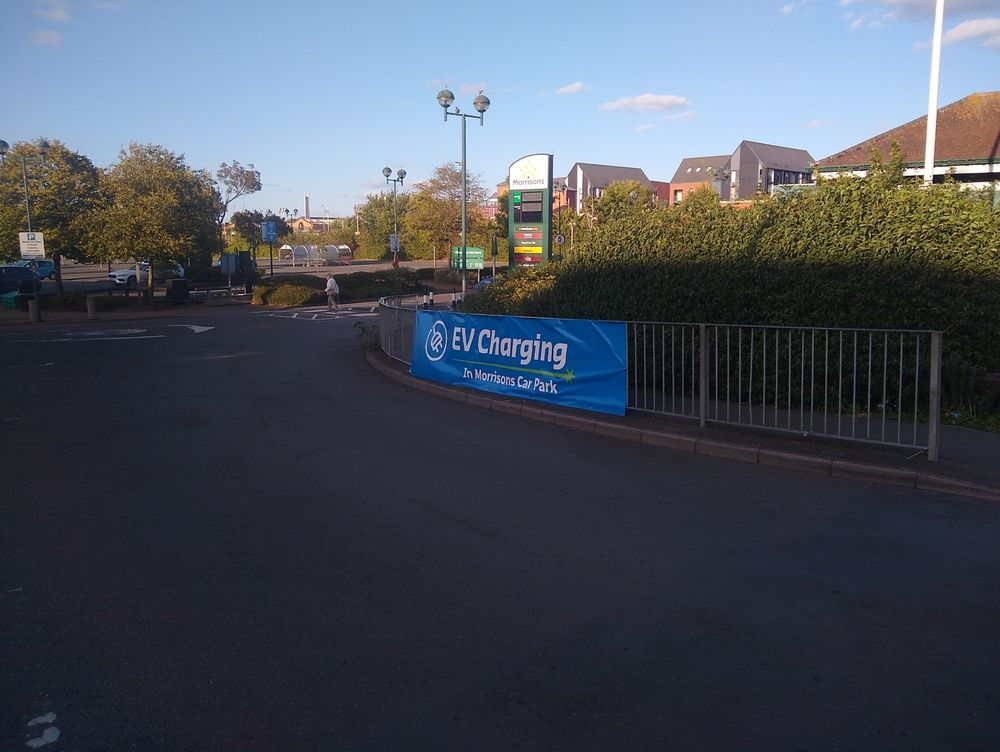 Large "EV Charging" banner on the fence alongside the entrance to Morrisons car park.