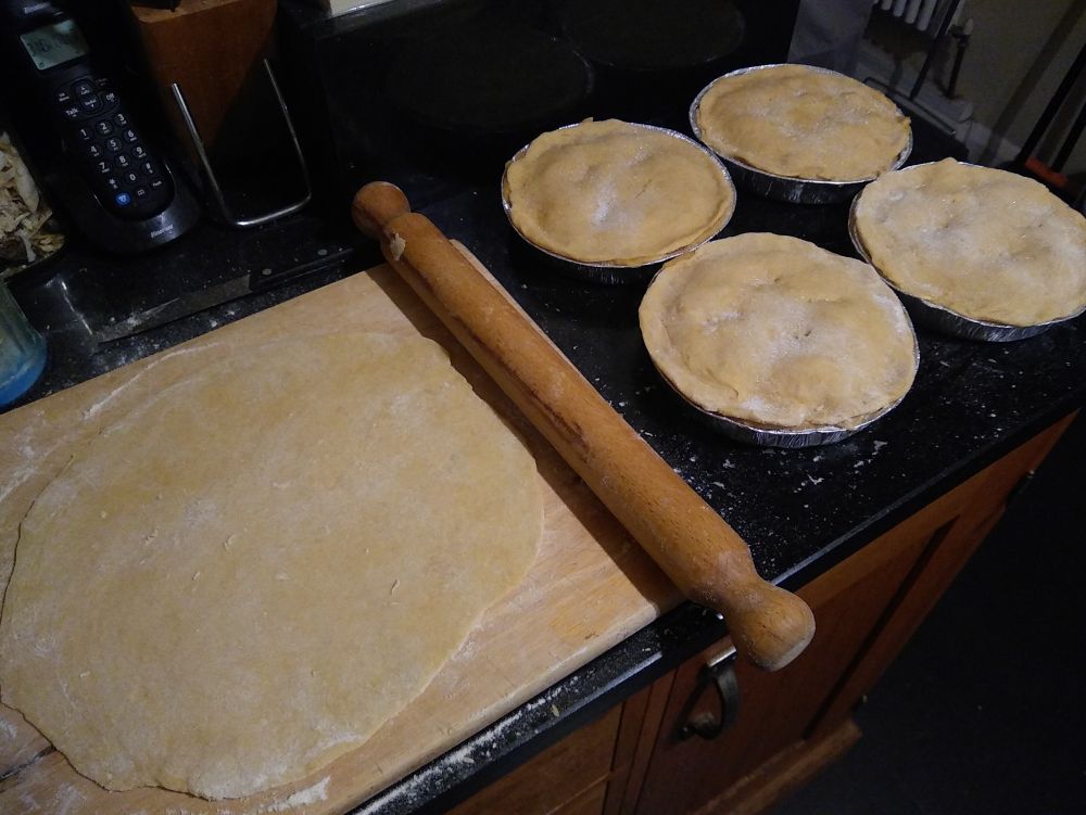 Four completed apple pies awaiting freezing and the base of another rolled out on a floured wooden board with a rolling pin next to it.