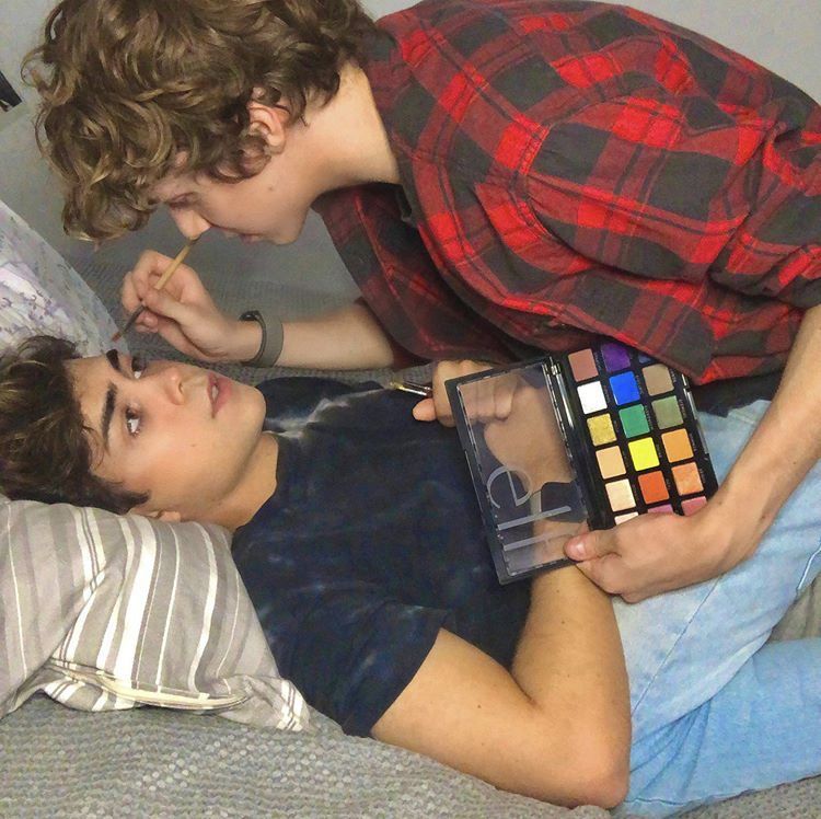 One person lying down while another person in a red and black plaid shirt leans over them holding a makeup palette.