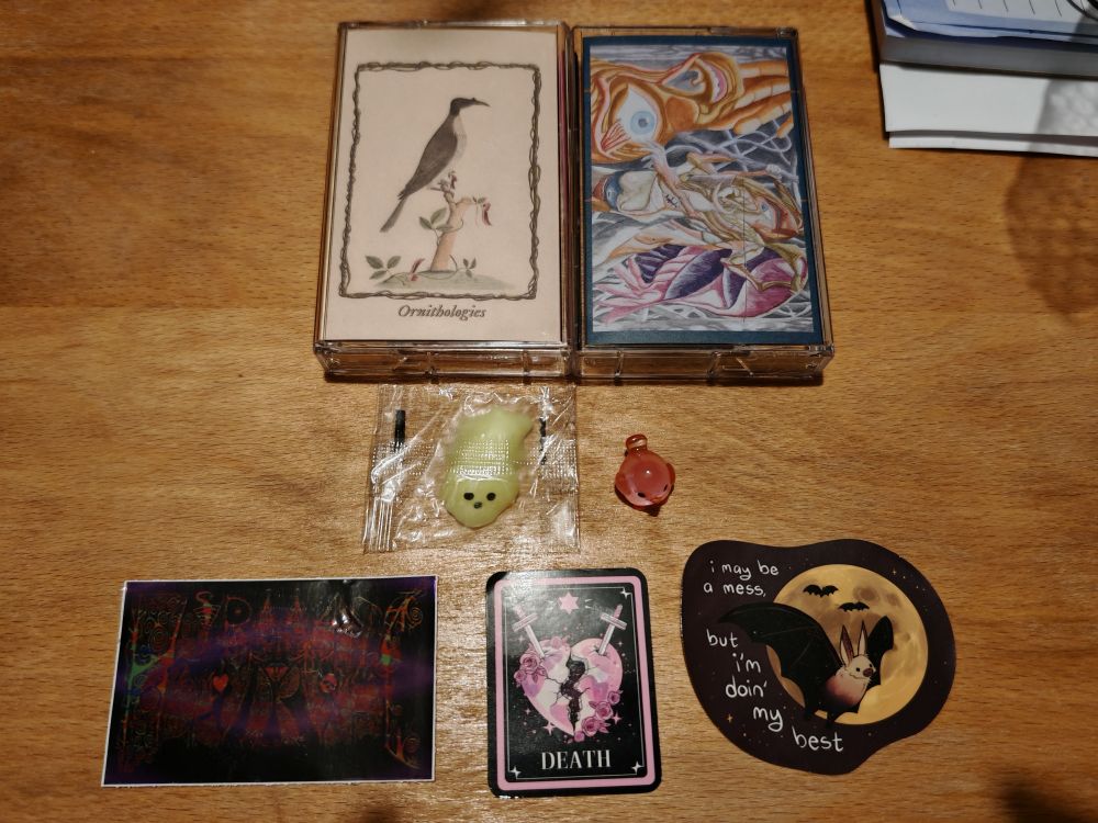 A cassette for Friarbird - Ornithologies next to a cassette for Hypomanic Daydream - The Yearning.

Below them are a little squishy green blob with eyes and nose in plastic packaging next to a tiny little rose glass bird.

Below are three stickers: one that says Hypomanic in very stylised and dark psychedelic colouring, one with a pink and white heart 'shell' that is splitting that has one side with cross-shaped gravestones sticking out that looks like daggers stabbing into it, labelled Death, and one that has a cartoon bat in front of a yellow moon with the caption "I may be a mess, but I'm doing my best"