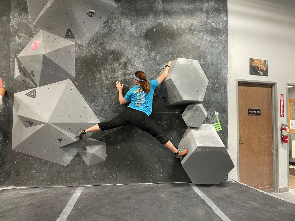 Rachelle is a few feet off the ground, on a challenging competition-style boulder set, facing the wall, legs spread out wide with her toes planted on two large volumes. Her right hand is gripping another large volume above her head, and her left palm is flat on the wall. Her head is turned left, looking ahead to her next movement.