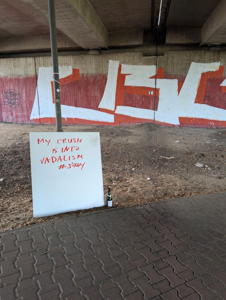 Ein weißes Schild mit der roten Aufschrift "MY CRUSH IS INTO VADALISM #SADLY" steht unter einer Brücke neben einer Sektflasche. Im Hintergrund ist eine Betonwand mit Graffiti.

