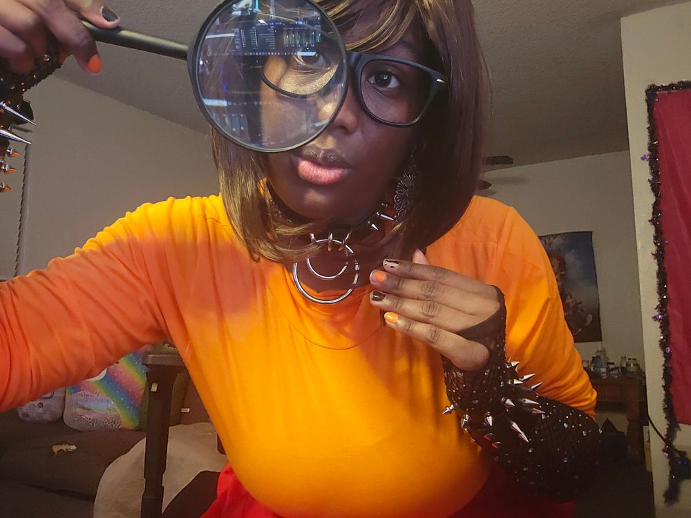 It's FlambéPlays cosplaying as Velma. She's looking through a magnifying glass, and she has on spiked wrist cuffs. Her spiked collar is around her neck. FlambéPlays nails are painted black and orange. 