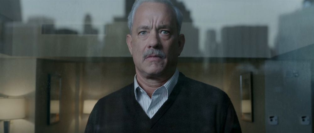 Tom Hanks as US Air Captain Chesley Sullenberger in Sully, white-grey hair and mustache, blue striped white dress shirt under dark v-neck sweater, behind glass window of what looks like a hotel room or meeting room, reflection of overcast sky in a city in the glass.