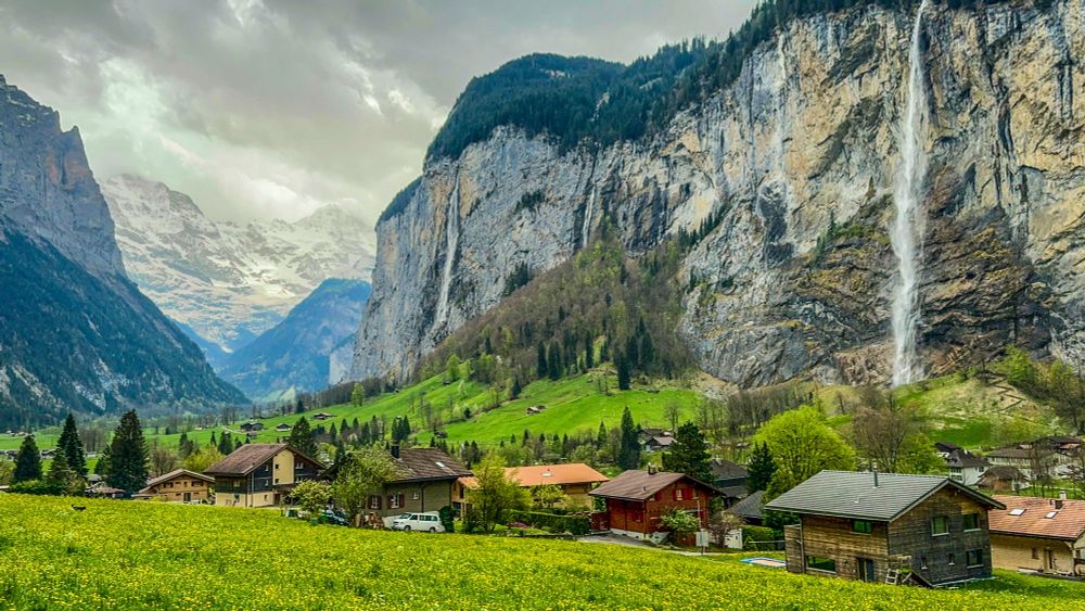 Verdant valley with a large cliff on the right side of the photo. A narrow waterfall falls from the cliff to the valley. A few typical Swiss houses are on the valley floor. 