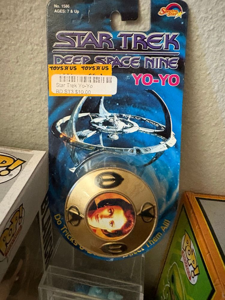 A Star Trek Deep Space Nine Yo-Yo in the package featuring Major Kira Nerys. The Yo-Yo is gold with a photo of Kira in the center with black Starfleet Delta and Bajoran symbols around it