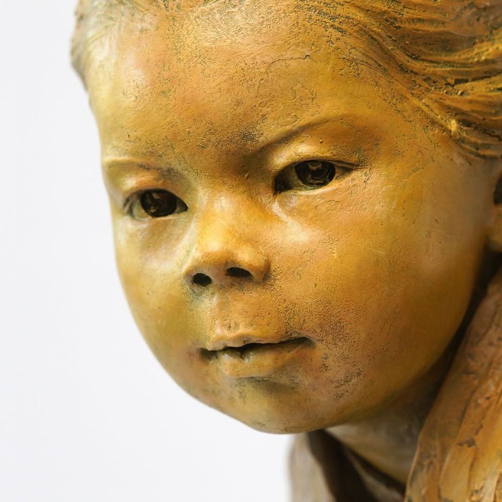 Detailed close-up of the face of artist Luo Li Rong's Bronze sculpture "Gaze of Childhood", which depicts a crouching child wearing a robe. "This sculpture emphasizes the child’s focused expression, aiming to convey the natural, quiet, and attentive moments of childhood. The posture and facial expression of the figure reflect the subtle interaction between the child and their surroundings, as if exploring the world around them, with a touch of innocence and curiosity"