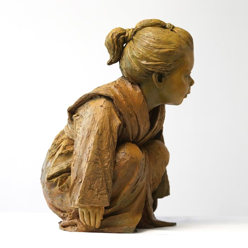 Side angle of artist Luo Li Rong's Bronze sculpture "Gaze of Childhood", which depicts a crouching child wearing a robe. "This sculpture emphasizes the child’s focused expression, aiming to convey the natural, quiet, and attentive moments of childhood. The posture and facial expression of the figure reflect the subtle interaction between the child and their surroundings, as if exploring the world around them, with a touch of innocence and curiosity"