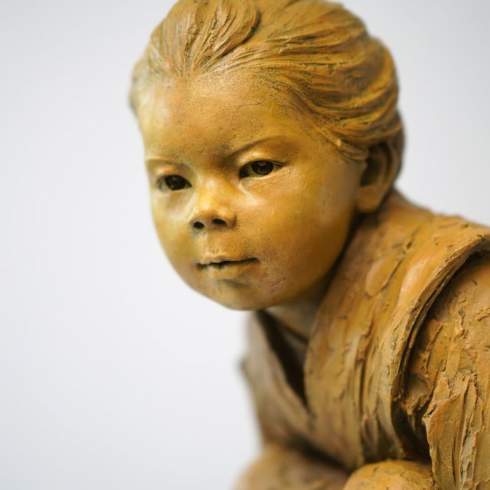 Detailed closeup on the face of Luo Li Rong's Bronze sculpture "Gaze of Childhood", which depicts a crouching child wearing a robe. "This sculpture emphasizes the child’s focused expression, aiming to convey the natural, quiet, and attentive moments of childhood. The posture and facial expression of the figure reflect the subtle interaction between the child and their surroundings, as if exploring the world around them, with a touch of innocence and curiosity"