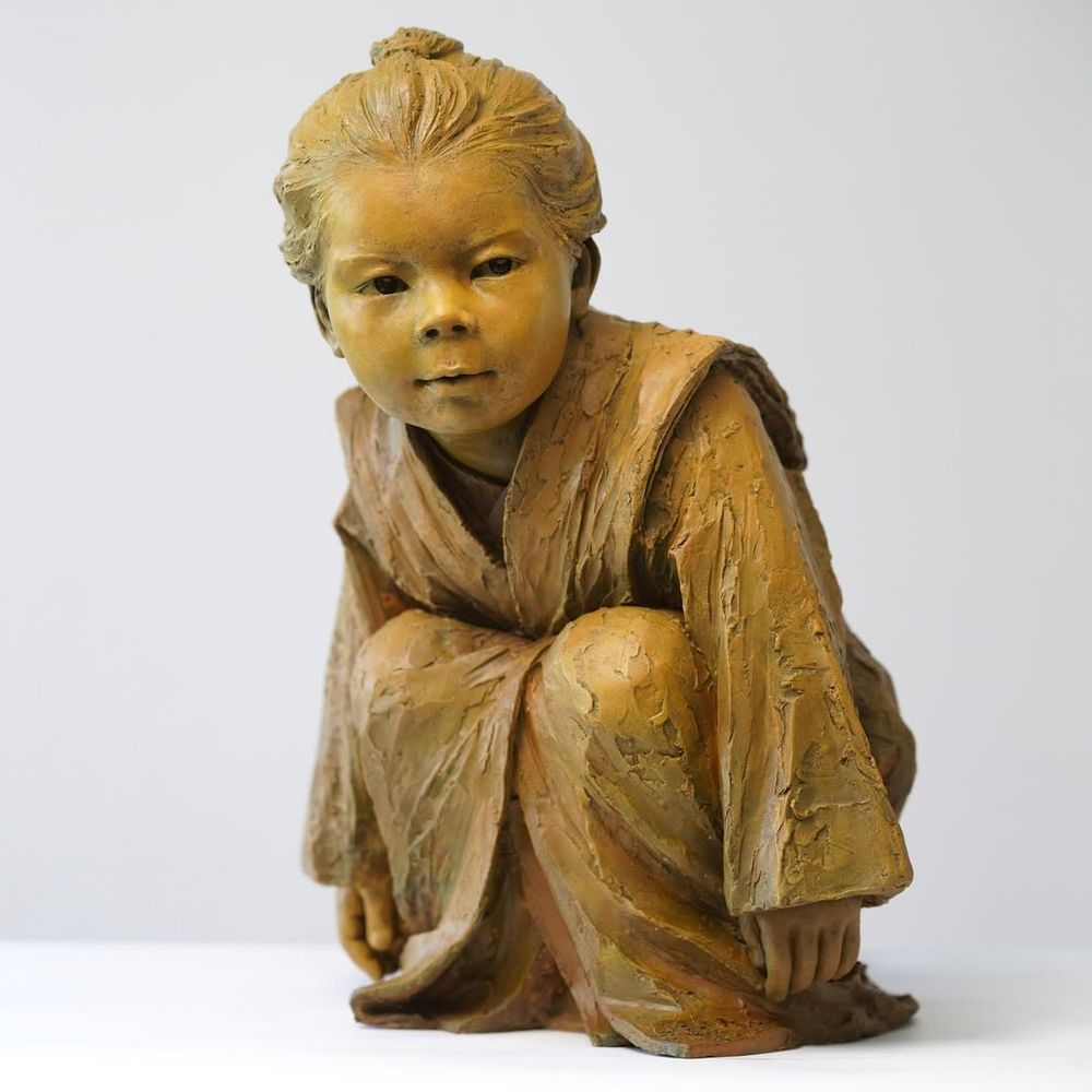 Bronze scuplture of a crouching child wearing a robe. "This sculpture emphasizes the child’s focused expression, aiming to convey the natural, quiet, and attentive moments of childhood. The posture and facial expression of the figure reflect the subtle interaction between the child and their surroundings, as if exploring the world around them, with a touch of innocence and curiosity"