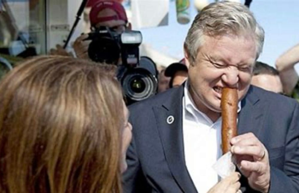 Photo of a middle aged white man, Marcus Bachmann, trying to eat a corn dog. He holds the dog with both hands and, with eyes closed, takes a tentative nibble. As a gay conversion practitioner, he seems to be having trouble opening up to take in the full experience. His wife Michelle, the brown haired head in the lower right, encourages him: “They’re really not that bad!”