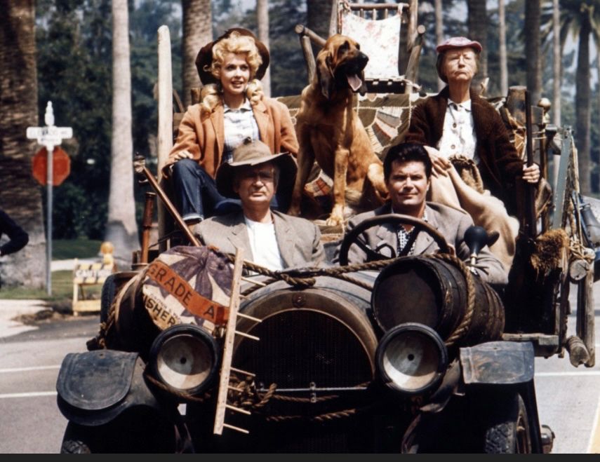 Photo from the tv series ‘The Beverley Hillbillies”. Uncle Jed and Jethro are in the front seat driving an open top, ramshackle vehicle with all their possessions, including a whiskey keg and a rocking chair, strapped to it. Elly May and Granny are sitting in the back with Duke, the bloodhound between them