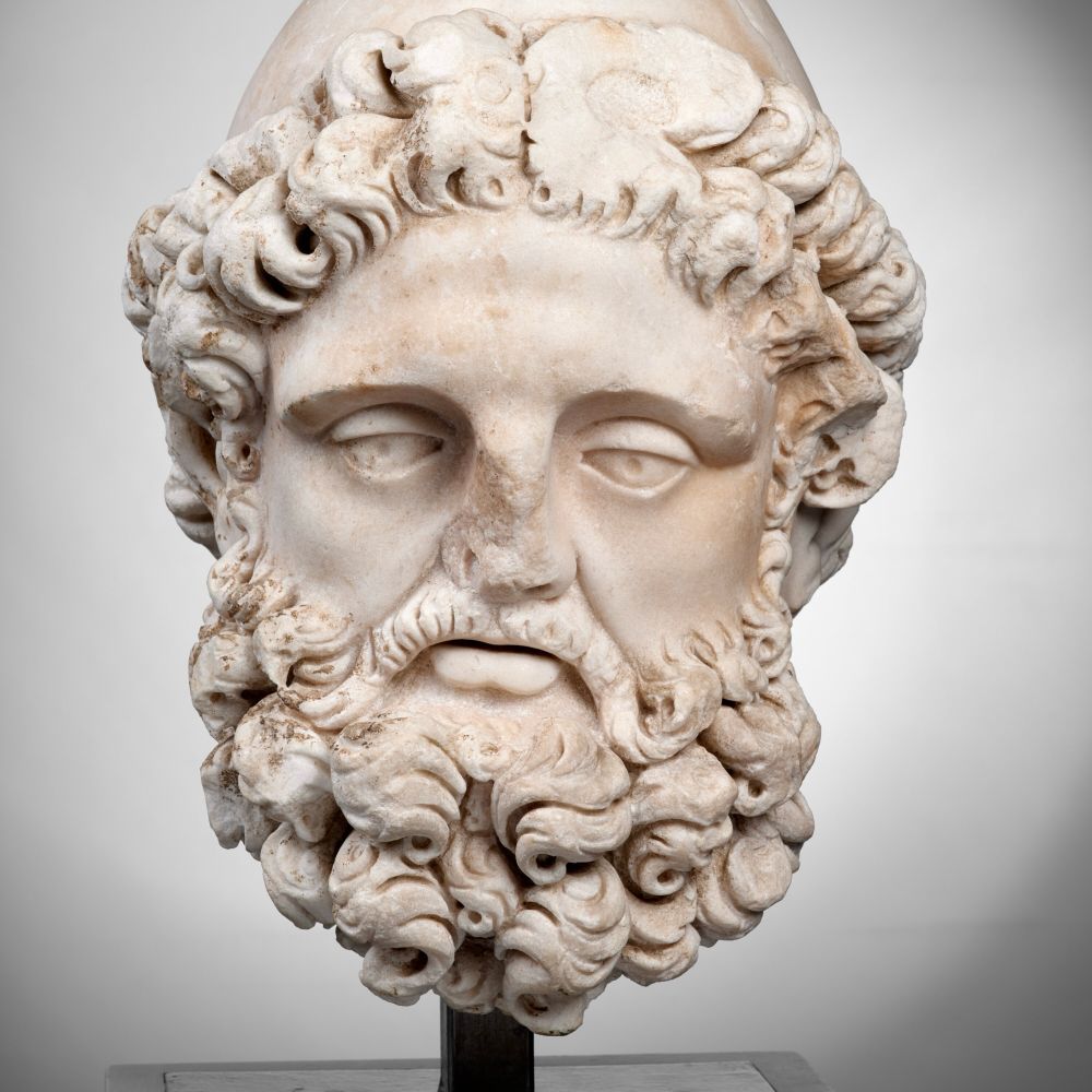 La même tête vue de face. La barbe est constituée de longues et grosses boucles. Une moustache fournie longe la lèvre supérieure pour se fondre dans la barbe.
Le nez est endommagé. 