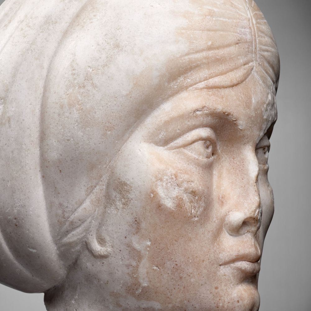 Détail d’un portrait féminin, vu de profil. La coiffure est composé d’un voile formant un turban autour. Le nez est endommagé. Les lèvres pincées. Les pommettes sont particulièrement saillantes. 