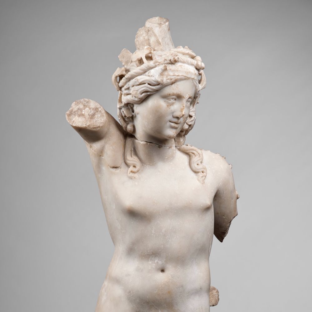 Partie supérieure d’une statuette en marbre ressentant Bacchus. Les deux bras sont brisés. Song bras droit était levé. Sa tête est légèrement tournée vers sa gauche. Ses cheveux longs retombent sur ses épaules. Il porte une couronne sur la tête. 