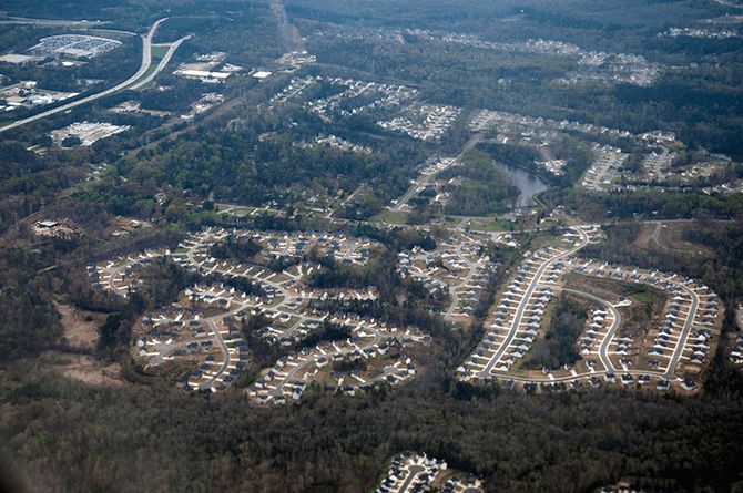 Aerial view of suburban tract homes in curving lines against pine trees, with an interstate highway in view.