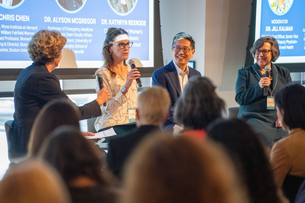 A panel of four people in front of a room with slides saying their names behind them. One person is looking at the other three. The second woman from the left is holding a mic. The other two are looking on smiling.