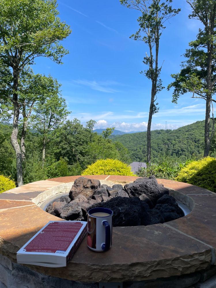 Wide blue sky over green mountains with a dormant fire pit, a book, and a cup of coffee in the foreground.