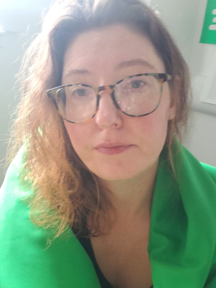 A white woman of around 40 wearing glasses and looking glum. She has a green sheet draped over her shoulders