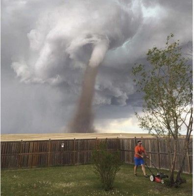 A 2017 photo of a Canadian man mowing grass in fenced back yard while a tornado touches down on the distant prairie in the background.