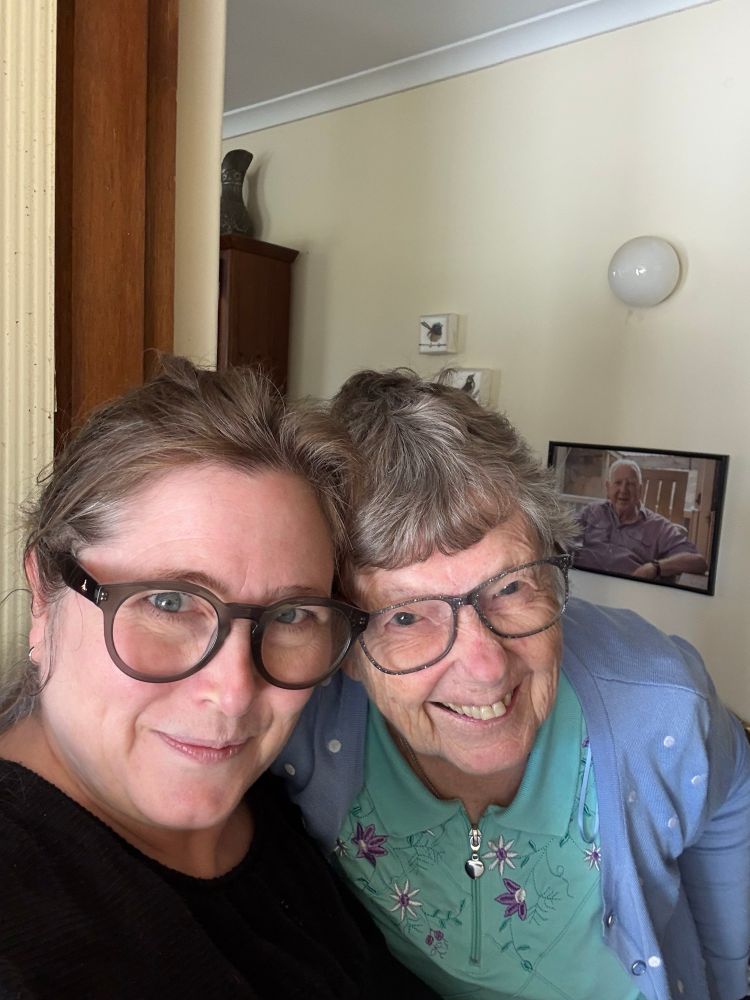 Liz is a middle aged woman with light brown hair pulled back and grey glasses. Her head is close to her Mum, an older lady with short grey hair. Both have happy smiles. Behind them is a large photo of Liz's Dad. 