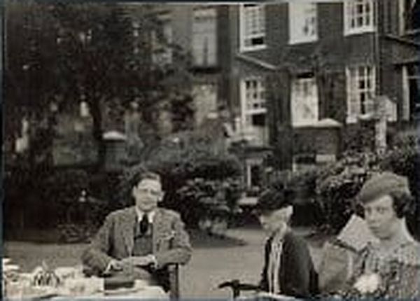 T.S. Eliot with his sister and his cousin

by Lady Ottoline Morrell
vintage snapshot print, 1934

The photograph is a vintage black-and-white snapshot from 1934, taken outdoors in what appears to be a garden or courtyard with tall brick buildings in the background. Three people sit at a table set for tea.

On the left, Eliot in a suit sits facing the camera, hands resting on the table. In the center, Eliot's cousin wearing a dark coat and hat sits slightly turned to the side, holding a cane. On the right, Eliot's sister in a patterned dress looks downward, her expression quiet and reserved.

https://en.wikipedia.org/wiki/T._S._Eliot#/media/File:Thomas_Stearns_Eliot_by_Lady_Ottoline_Morrell_(1934).jpg