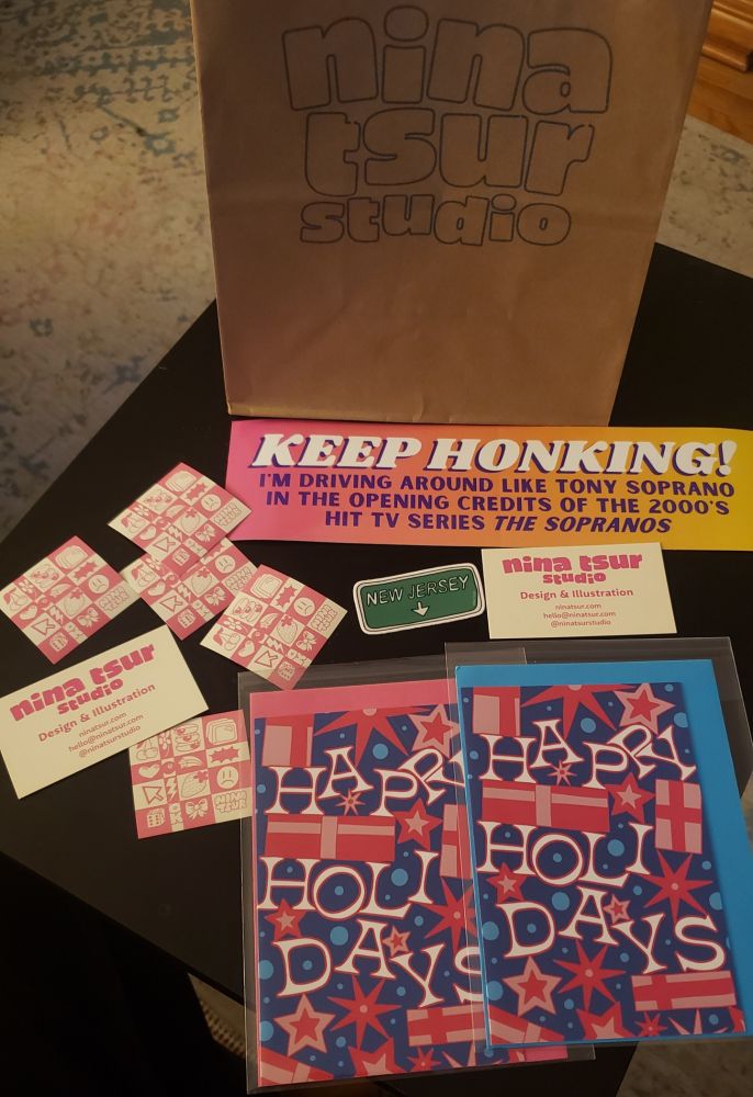 Two holiday cards, some stickers (pink ones and a New Jersey one in green), a few business cards, and an amazing bumper sticker mentioning the Sopranos. They are presented on a small and wooden TV tray.