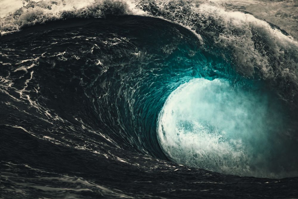 A dramatic close-up of a massive ocean wave curling into a hollow tube, with frothy white water cresting at the top and deep turquoise hues illuminated in the center of the barrel. The surrounding water appears dark and turbulent, contrasting with the bright, glowing interior of the wave.