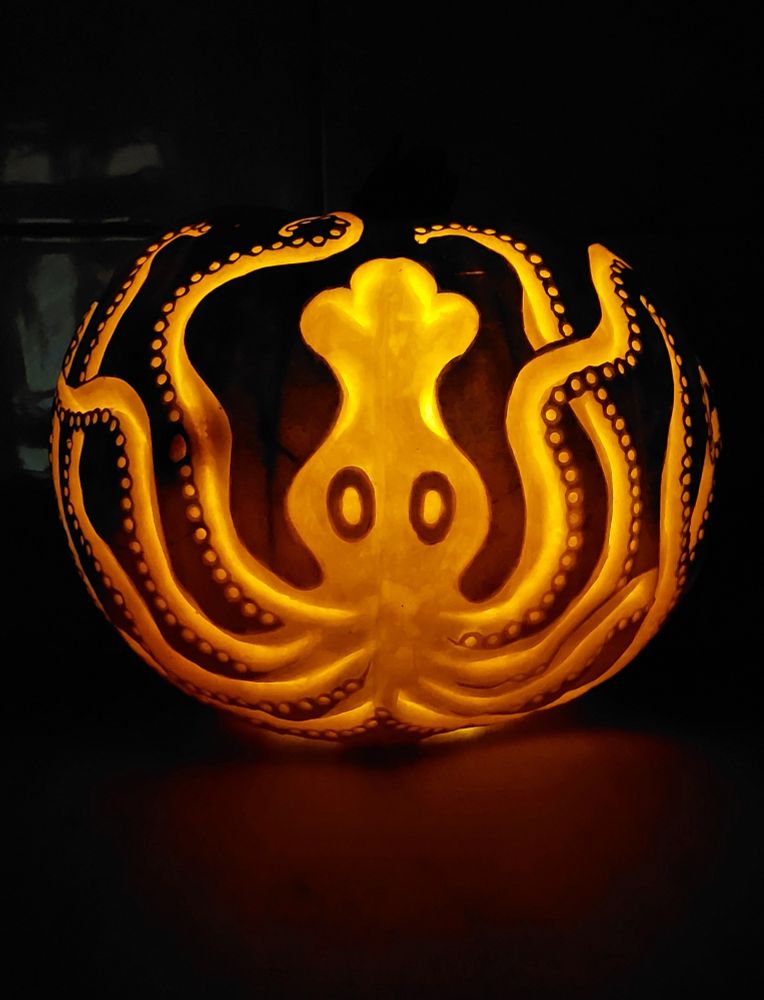 A jack o lantern, lit up orange, in the dark. It's been carved with a Minoan style octopus, so it's simplified and a little goofy looking. It has a big bulbous head, two oval eyes, and eight tentacles weaving up from the bottom of the body and the pumpkin. The tentacles all have a series of small holes for suckers along the bottom of their length.