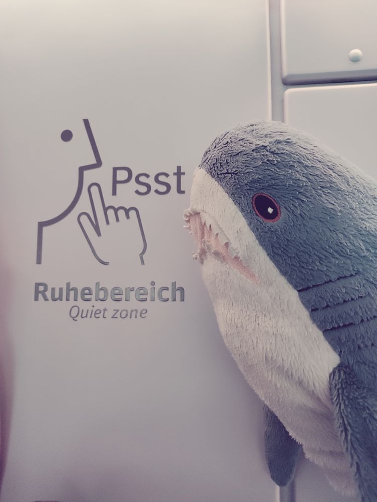 Symbol und Schriftzug an einer weißen Wand in einem Zug der Deutschen Bahn: "Psst ruhebereich - Quiet Zone". Von rechts schaut ein kleiner Stoffhai von Ikea den Schriftzug an. Er hat ein wachsames Auge Richtung Kamera, sodass er auf jeden Fall sieht, wer es war, wenn jemand Lärm macht
