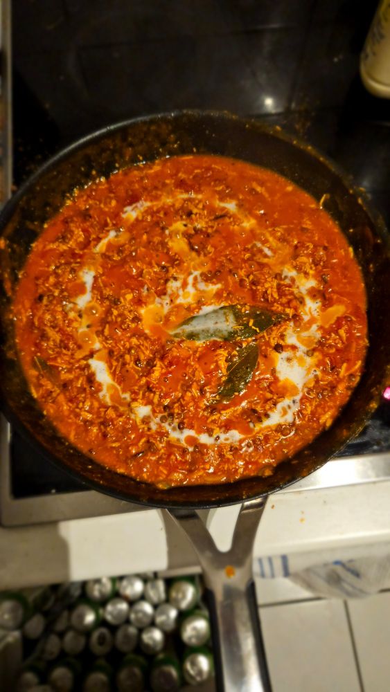 Pfanne auf dem Herd. In der Pfanne befindet sich Curry ähnliches essen. Man sieht insbesondere geriebenen Tofu & linsen in einer roten Tomatensauce. In der Mitte schwimmen 2 Lorbeerblätter und mit hafersahne ist eine Spirale reingezogen. 