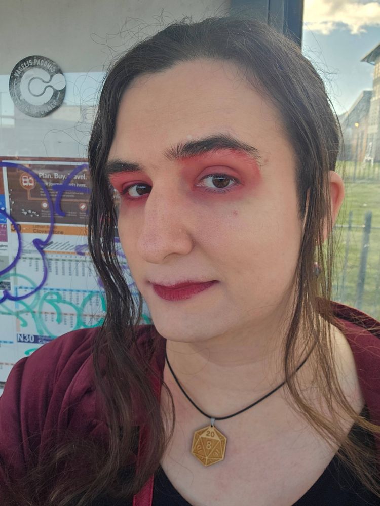 Selfie of me, a tall white woman with wavy brown hair standing in front of a defaced bus stop. I have rather garish bloody style red lipstick and eyeshadow on, as well as a woodcut pendant of a D20 and an oxblood red jacket. I'm smiling simply at the camera.
