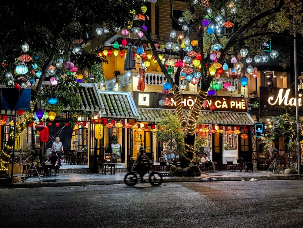 We are standing in a darkened, evening street watching the silhouette of a child ride past a brightly decorated cafe in eastern Hanoi. The cafe is painted the distinctive ocre-gold of many old buildings in the city (though it is quite new, in this case) and has an overhanging front set of eaves meant to resemble the tiled roofs of classical Vietnamese structures. A pair of trees on the side walk out front are brightly lit. Fairy lights wind around the trucks in dense configurations, while the upper branches are filled with multi-colored lanterns. A pair of men sit at a table in the shadows on the left while another sits alone on the sidewalk to the right. 