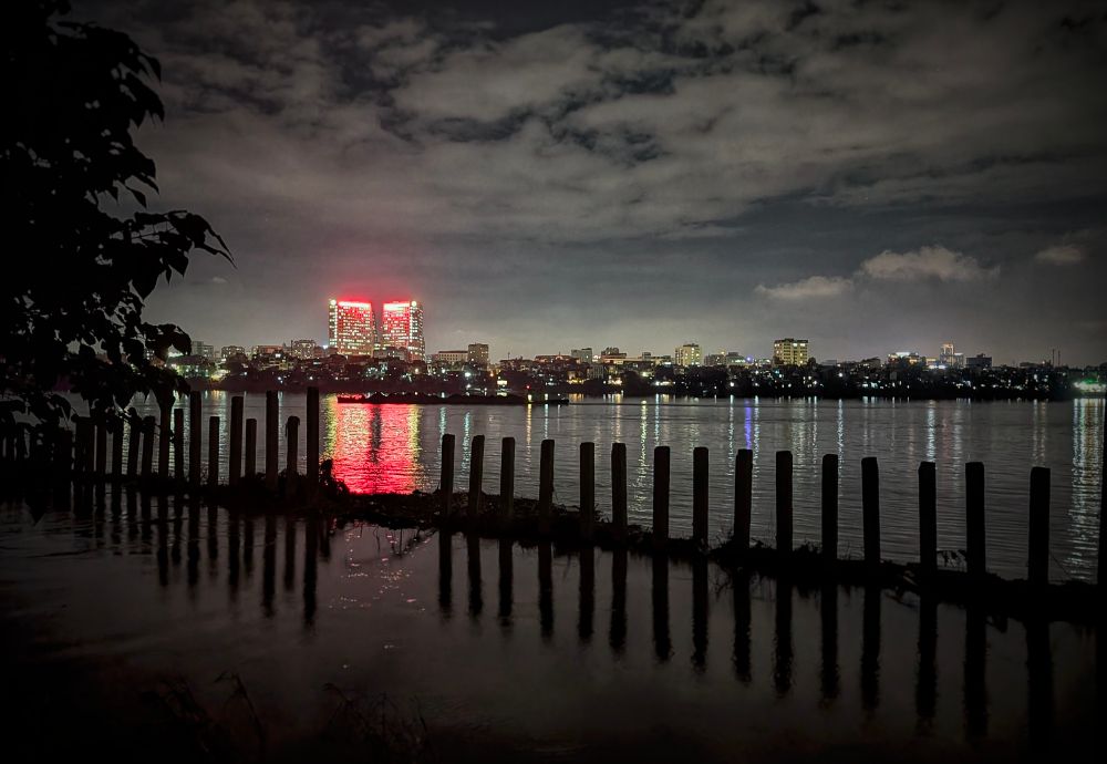 An evening photo of the Red River from the banks of Lông Biên in Hanoi. From where we stand, we see a row of river pilings extending diagonally out into the water. The dark silhouette of a tree frames the left side of the image. Across the wide expanse of the river we see a relatively low-rise skyline of illuminated houses and buildings lining the river with a few larger structures in the faint background. Standing brightly above the scene, however, is the 108 Army Hospital. Its luminous red sign reflects off the river surface. The city lights reflect off splotchy, low clouds. 
