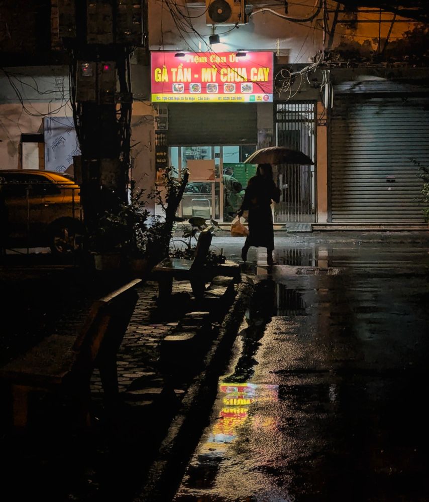 A night photo down a rain-soaked alleyway behind an apartment block in the Nghĩa Tân Ward of Hanoi. We are standing on the edge of a small sân chơi that backs an old khrushchevka-style apartment building. A small side street winds its way around the concrete pad of this recreation space as rain water collects by the curb. We can see the outlines of concrete benches and the reflection of a half-shuttered noodle joint at the end of the street. Its still-illuminated sign glows red with yellow text. In clear silhouette, there strides a woman holding an umbrella in one hand and a plastic bag in the other stepping lightly between puddles. 