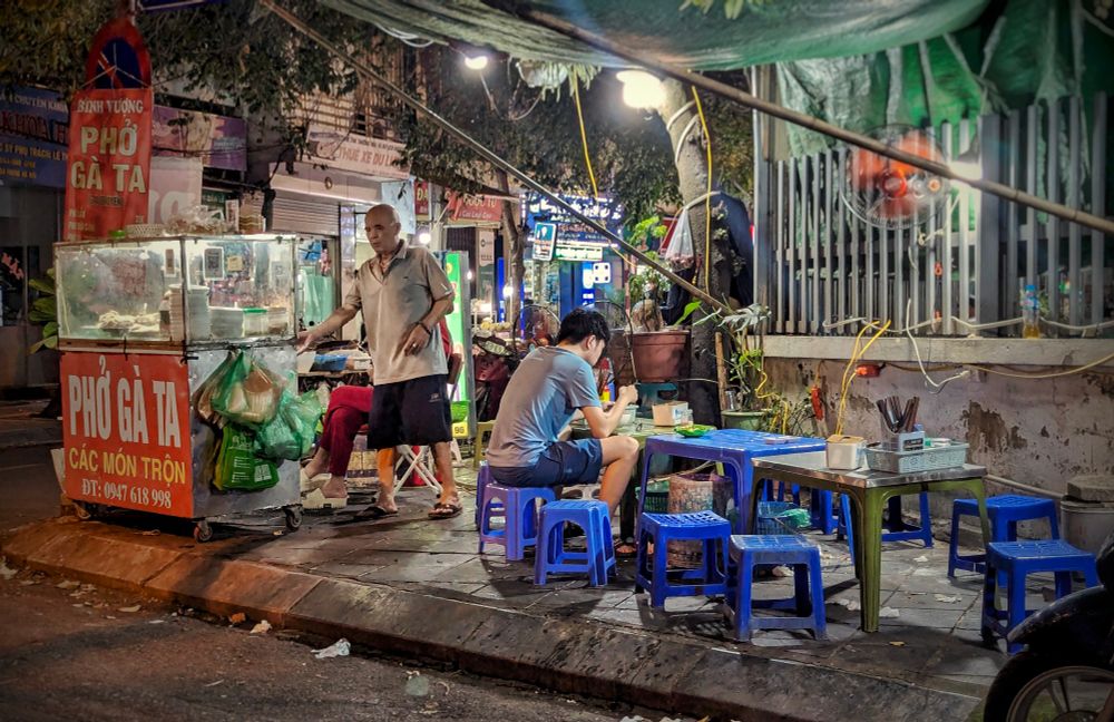 An open air chicken phở stall on a sidewalk in southern Hanoi. A seating area of blue and green plastic tables and stools sits to the right under a makeshift awning of green plastic held up by bamboo pokes fastened to the adjacent wall. A single patron in a light blue t-shirt and dark blue shorts sits over a bowl of noodles. On the left, a bald man in a light, short sleeve collared shirt and dark shorts stands beside the stall while a woman in matching red velour pants and shirt just just behind him. The stall sign is red with white and yellow lettering facing the street. A banner hangs from an adjacent tree with a similar color scheme and message. The frontages of mostly closed businesses can be seen in the background along a small commercial street. 