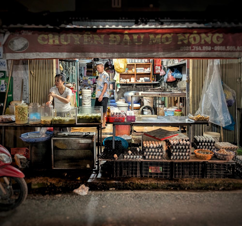 A man and a woman tend to their wares in a small, back-alley shop in Hanoi. The woman, in a white shirtsleeve shirt and black-and-white check pants stands behind a row of large receptacles with what appear to be various fermented vegetables waiting to be bagged and taken home. On the right are stacks of eggs in 6x6 foam flats. All sits under a red canopy with yellow lettering. In the background we see a man in a floral-print shirt and black shorts. He has close cropped hair and moves among shelves of ingredients, packaging, and utensils. 