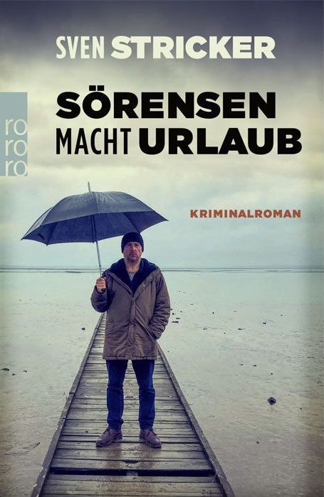 The image shows the book cover for "Sörensen Macht Urlaub" by Sven Stricker. The book is likely a crime novel (Kriminalroman). The cover features a man standing on a wooden pier, holding a black umbrella. He's dressed in a dark hat, a tan jacket, and jeans. The setting is a beach, with a dull sky in the background. The color scheme of the cover is muted, creating a slightly gloomy atmosphere.