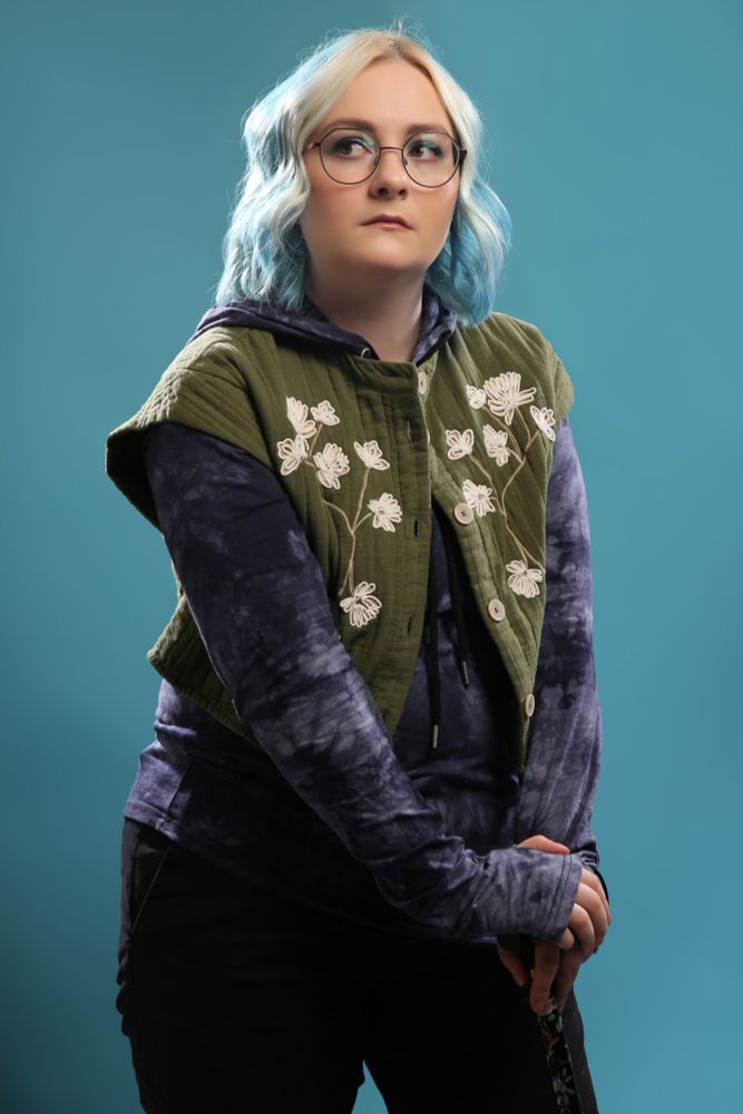 A nonbinary person standing with both hands leaning on a cane, looking away in a thoughtful pose. They are white, and have blue, wavy hair with white strips at the front, and round framed glasses. They are wearing a long-sleeved hoodie in a dappled blue pattern, layered with a cropped green cotton jacket with embroidered white flowers. The background is turquoise.