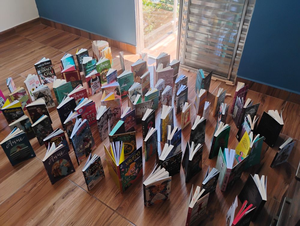 foto de vários livros e mangás em pé e abertos.
eles estão em um chão marrom que imita madeira e estão de frente para uma porta de metal aberta que leva para a sacada e entra um pouco de luz do sol.