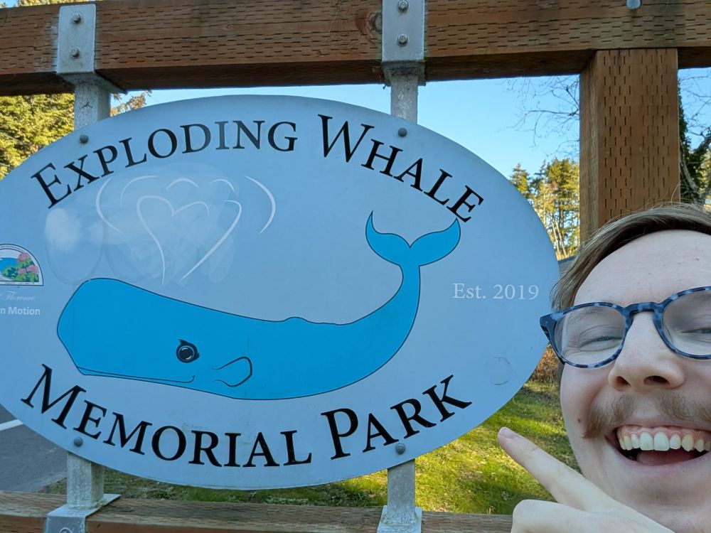 Roman pointing at a sign with a cartoon sperm whale shooting water and the words "Exploding Whale Memorial Park. Est. 2019"