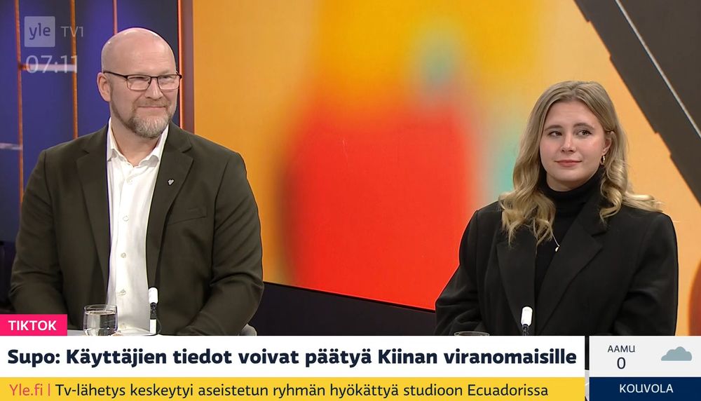Mikko Kiviharju sitting on the left, smiling and dressed in a brown jacket and white shirt, Laura Kankaala sitting on the right, dressed in black. Red and yellow studio backdrop.