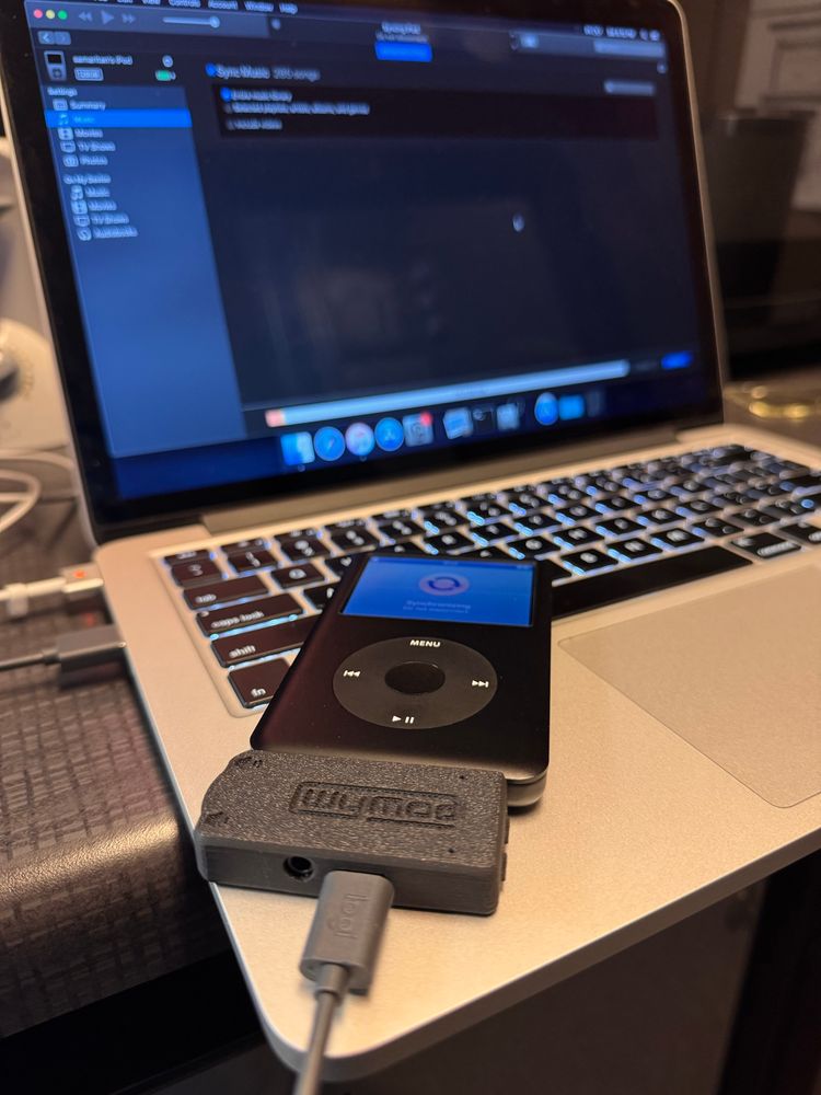 An iPod classic 7th generation plugged into a MacBook Pro with a USB C adapter with built in Bluetooth and FireWire, the name Wymod inscribed on the top of the adapter. 